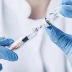 Ręce w niebieskich rękawiczkach przygotowują zastrzyk z biofuroksymem, pobierając płyn ze szklanej fiolki do strzykawki.