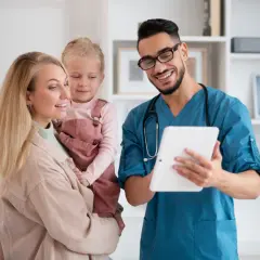 Lekarz w niebieskim kitlu pokazuje coś na tablecie uśmiechniętej kobiecie trzymającej dziecko.