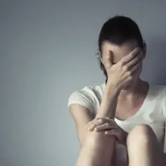 Kobieta w białej koszulce siedzi przy szarej ścianie, zakrywając twarz dłońmi. Cierpi, jakby szukała ukojenia, może po trudnym dniu, który przypominał lerivon.