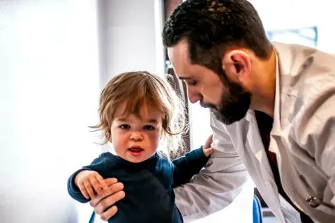 Lekarz wspiera małe dziecko, które ma achondroplazję. Dziecko patrzy w kamerę, a lekarz pochyla się nad nim.