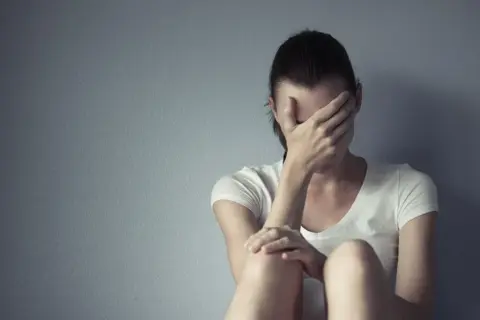 Kobieta w białej koszulce siedzi przy szarej ścianie, zakrywając twarz dłońmi. Cierpi, jakby szukała ukojenia, może po trudnym dniu, który przypominał lerivon.