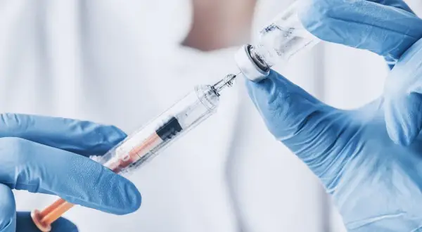 Ręce w niebieskich rękawiczkach przygotowują zastrzyk z biofuroksymem, pobierając płyn ze szklanej fiolki do strzykawki.