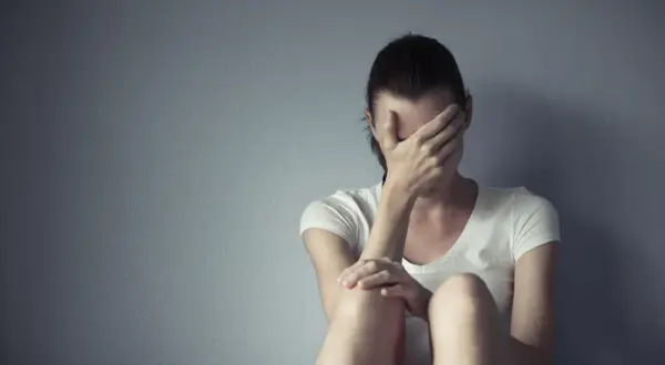 Kobieta w białej koszulce siedzi przy szarej ścianie, zakrywając twarz dłońmi. Cierpi, jakby szukała ukojenia, może po trudnym dniu, który przypominał lerivon.