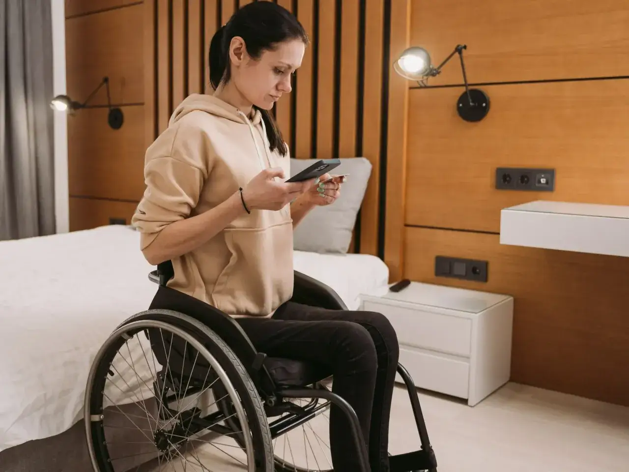 Kobieta na wózku inwalidzkim, zmagająca się z miopatią, korzysta z telefonu i karty płatniczej w hotelowym pokoju.