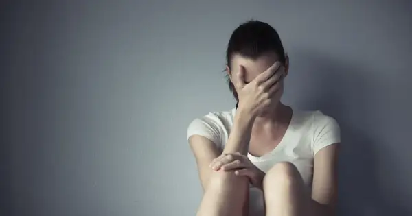 Kobieta w białej koszulce siedzi przy szarej ścianie, zakrywając twarz dłońmi. Cierpi, jakby szukała ukojenia, może po trudnym dniu, który przypominał lerivon.